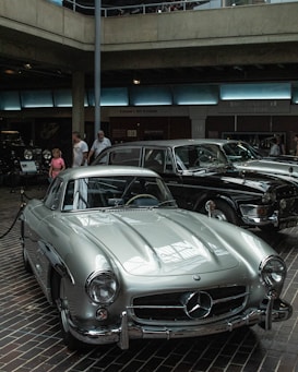 A classic silver Mercedes-Benz car is prominently displayed indoors, reflecting the surrounding lights. Nearby, a black vintage car is visible, with a couple of people walking by in casual attire. The setting appears to be an automotive museum or exhibition with a modern architectural design featuring concrete and glass elements.