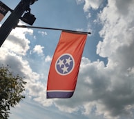 A red flag with three central white stars inside a blue circle, beneath a sky with fluffy white clouds and a hint of sunlight. The flag appears to be mounted on a black pole, and some green tree leaves are visible in the corner.