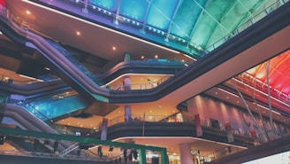 An interior view of a modern shopping mall with multiple levels connected by escalators. The ceiling features dynamic lighting in gradient colors ranging from blue to orange. Architectural elements display sleek lines and a contemporary design with glass and metal materials.