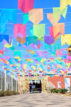 Colorful paper banners strung above a walkway create a festive and vibrant atmosphere. The banners are arranged in rows, with hues of pink, blue, yellow, green, and purple contrasting against a clear blue sky. The pathway leads to a building entrance adorned with decorations and natural greenery.
