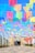 Colorful paper banners strung above a walkway create a festive and vibrant atmosphere. The banners are arranged in rows, with hues of pink, blue, yellow, green, and purple contrasting against a clear blue sky. The pathway leads to a building entrance adorned with decorations and natural greenery.