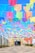 Colorful paper banners strung above a walkway create a festive and vibrant atmosphere. The banners are arranged in rows, with hues of pink, blue, yellow, green, and purple contrasting against a clear blue sky. The pathway leads to a building entrance adorned with decorations and natural greenery.