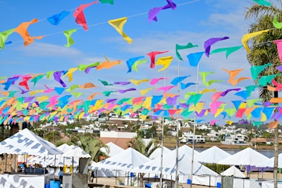Colorful flags made of fabric, in shades of blue, yellow, orange, green, and purple, are strung across a blue sky, creating a festive atmosphere. Below, a row of white tents is set up in an outdoor area, surrounded by palm trees and residential buildings in the background.
