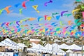 Colorful flags made of fabric, in shades of blue, yellow, orange, green, and purple, are strung across a blue sky, creating a festive atmosphere. Below, a row of white tents is set up in an outdoor area, surrounded by palm trees and residential buildings in the background.