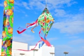 A colorful kite with floral patterns and vibrant ribbons flying in a clear blue sky. The background shows a building and several streetlights.
