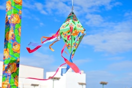 A colorful kite with floral patterns and vibrant ribbons flying in a clear blue sky. The background shows a building and several streetlights.