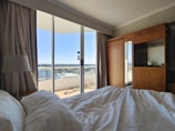 A cozy hotel room with a rumpled bed in the foreground. There is a large window displaying an outdoor balcony with a view of the cityscape. The room contains wooden furniture, including a wardrobe and a cabinet with a lamp. Light curtains frame the window, and there is a chair visible on the balcony.