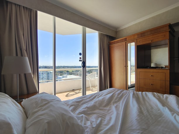 A cozy hotel room with a rumpled bed in the foreground. There is a large window displaying an outdoor balcony with a view of the cityscape. The room contains wooden furniture, including a wardrobe and a cabinet with a lamp. Light curtains frame the window, and there is a chair visible on the balcony.