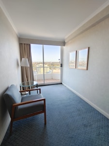 A small, narrow room with light-colored walls features a simple setup that includes a single armchair with wooden arms, a small round glass table, a floor lamp, and two framed pictures on the wall. Large glass sliding doors at the end of the room lead to a balcony overlooking a cityscape.