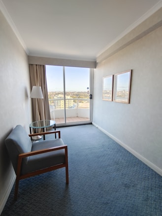 A small, narrow room with light-colored walls features a simple setup that includes a single armchair with wooden arms, a small round glass table, a floor lamp, and two framed pictures on the wall. Large glass sliding doors at the end of the room lead to a balcony overlooking a cityscape.