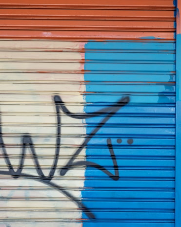A wall with a metal roller shutter painted in three sections: orange on top, beige in the center, and blue on the right. A black graffiti outline resembling abstract or stylized shapes extends across the surface.