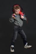 A young child stands confidently, wearing headphones and holding colorful toy guns. The child is dressed in a patterned shirt, dark jeans, and sneakers. The background is a dark shade, highlighting the subject.