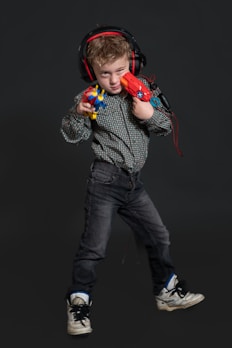 A young child stands confidently, wearing headphones and holding colorful toy guns. The child is dressed in a patterned shirt, dark jeans, and sneakers. The background is a dark shade, highlighting the subject.