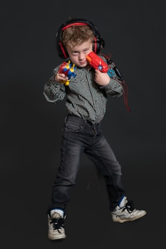 A young child stands confidently, wearing headphones and holding colorful toy guns. The child is dressed in a patterned shirt, dark jeans, and sneakers. The background is a dark shade, highlighting the subject.