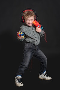 A young child is smiling broadly while wearing large headphones and holding a brightly colored toy gun and a toy character in each hand. The child is dressed in a patterned shirt and jeans, and appears to be in a playful and energetic pose.