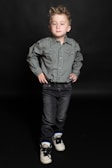 A young child stands confidently against a dark background, wearing a green checkered shirt and dark jeans. The child's hairstyle is spiky and the expression is calm yet self-assured. The shoes are white with black laces and blue accents.