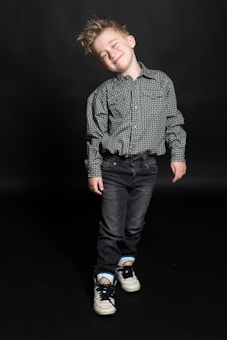 A young child with spiky hair is dressed in a checkered shirt, dark jeans, and white sneakers. The child is leaning slightly and smiling with closed eyes against a dark background.