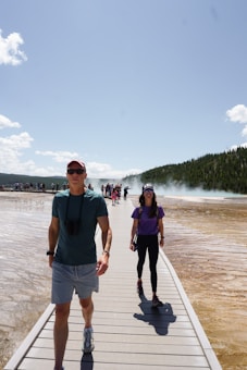 A boardwalk runs through a geothermal area with steam rising in the background. Two people walk toward the camera, enjoying a sunny day; one wears a purple shirt and black pants, while the other sports a green shirt and shorts. Other visitors can be seen further down the boardwalk, and lush trees line the horizon under a clear blue sky.