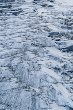 An aerial view of a glacier with numerous crevasses and rugged icy ridges. The surface is marked by streaks of dark sediment, creating intricate patterns against the white and bluish ice. The texture is rough and uneven, indicating the natural erosion and movement of the glacier over time.