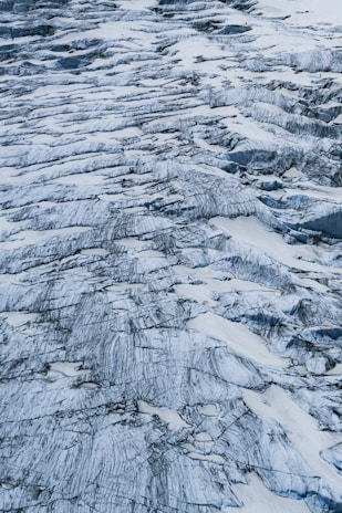 An aerial view of a glacier with numerous crevasses and rugged icy ridges. The surface is marked by streaks of dark sediment, creating intricate patterns against the white and bluish ice. The texture is rough and uneven, indicating the natural erosion and movement of the glacier over time.