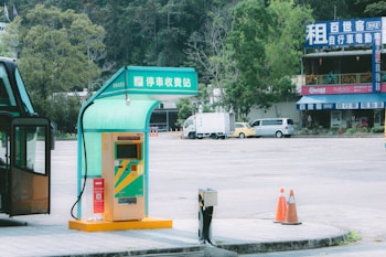 A parking payment station is situated under a shelter with a green and yellow color scheme. In the background, there are empty parking spaces, a few vehicles, and a building with signs and bicycles on the balcony. Trees add a natural element to the scene on the left side.