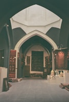 An arched interior space filled with a variety of colorful, patterned rugs displayed on the walls and laid out on the floor. Ceramic pots and other decorative items are showcased on a low table to the right, and framed artworks are stacked against the walls.
