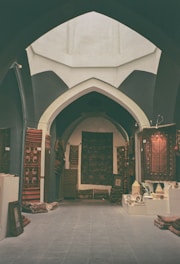 An arched interior space filled with a variety of colorful, patterned rugs displayed on the walls and laid out on the floor. Ceramic pots and other decorative items are showcased on a low table to the right, and framed artworks are stacked against the walls.