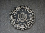 A circular seal of the Federal Bureau of Investigation is set against a textured surface. The seal includes the Department of Justice's name, an eagle, a shield, and the words Fidelity, Bravery, Integrity.