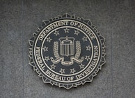 A circular seal of the Federal Bureau of Investigation is set against a textured surface. The seal includes the Department of Justice's name, an eagle, a shield, and the words Fidelity, Bravery, Integrity.