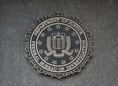 A circular seal of the Federal Bureau of Investigation is set against a textured surface. The seal includes the Department of Justice's name, an eagle, a shield, and the words Fidelity, Bravery, Integrity.