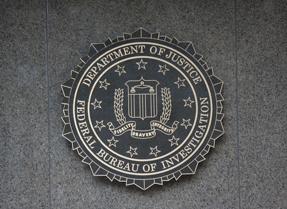 A circular seal of the Federal Bureau of Investigation is set against a textured surface. The seal includes the Department of Justice's name, an eagle, a shield, and the words Fidelity, Bravery, Integrity.