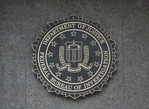 A circular seal of the Federal Bureau of Investigation is set against a textured surface. The seal includes the Department of Justice's name, an eagle, a shield, and the words Fidelity, Bravery, Integrity.
