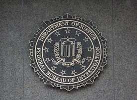 A circular seal of the Federal Bureau of Investigation is set against a textured surface. The seal includes the Department of Justice's name, an eagle, a shield, and the words Fidelity, Bravery, Integrity.