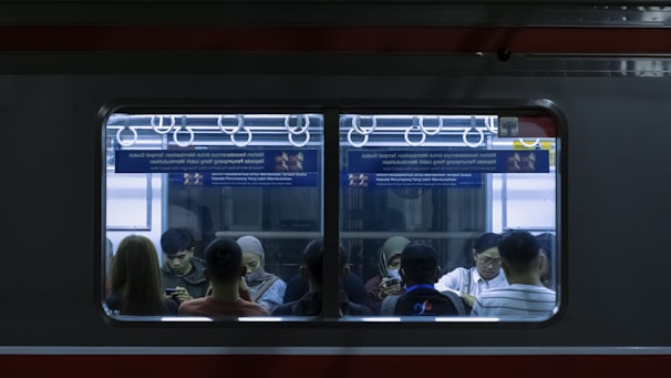 Inside view of a subway car where several people are seated, mostly absorbed in their phones. The interior features blue lighting and metal grab handles are visible above the passengers. Advertising posters are displayed above the windows.