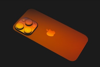 A smartphone with a sleek design, featuring a prominent brand logo centered on its back. The back of the device is highlighted by its three camera lenses arranged in an elevated square formation at the upper left corner. The surface has a reflective finish, capturing ambient light.
