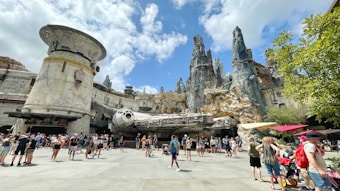 A theme park setting with a large spacecraft model surrounded by rocky, cliff-like structures. People are gathered around, some standing and others walking by. The architecture is futuristic with metallic and stone elements. The sky is partly cloudy, and there are trees and umbrellas providing shade.