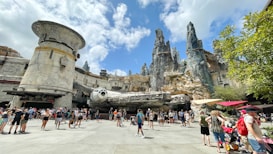 A theme park setting with a large spacecraft model surrounded by rocky, cliff-like structures. People are gathered around, some standing and others walking by. The architecture is futuristic with metallic and stone elements. The sky is partly cloudy, and there are trees and umbrellas providing shade.