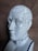 A ceramic bust depicting a phrenology head with labeled regions indicating different mental faculties and characteristics. The bust is adorned with a beaded necklace and set against a neutral, textured background.