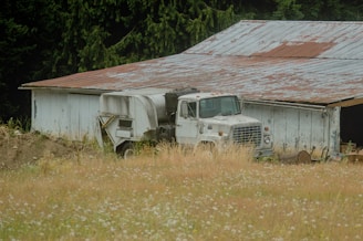 A rustic old truck parked beside a red barn with Idaho mountains rising in the background under a clear blue sky.