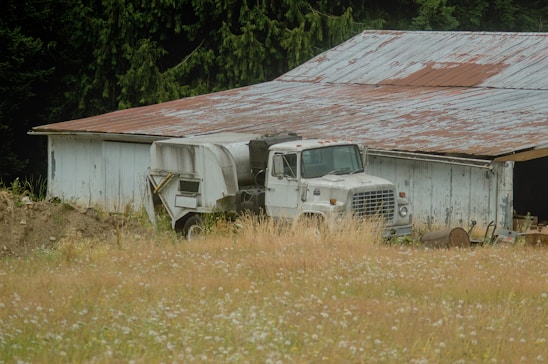 A rustic old truck parked beside a red barn with Idaho mountains rising in the background under a clear blue sky.