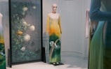 A mannequin wearing a long gradient dress transitioning from yellow at the top to green at the bottom, featuring a nature-themed design. The background displays a decorative panel with intricate botanical patterns, including large flowers and leaves.