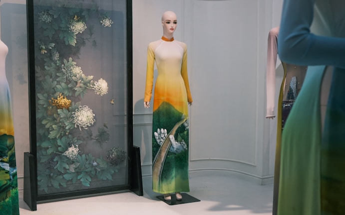 A mannequin wearing a long gradient dress transitioning from yellow at the top to green at the bottom, featuring a nature-themed design. The background displays a decorative panel with intricate botanical patterns, including large flowers and leaves.