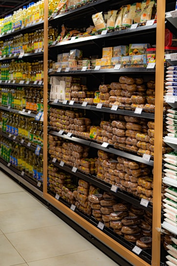 Shelves in a grocery store are stocked with various packaged food items. The left side features a range of cooking oils, while the right side displays a selection of baked goods or packaged snacks. Each shelf is orderly, with products neatly arranged in rows and labels visible for pricing.