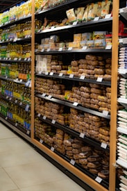 Shelves in a grocery store are stocked with various packaged food items. The left side features a range of cooking oils, while the right side displays a selection of baked goods or packaged snacks. Each shelf is orderly, with products neatly arranged in rows and labels visible for pricing.