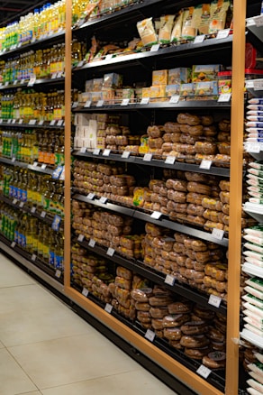 Shelves in a grocery store are stocked with various packaged food items. The left side features a range of cooking oils, while the right side displays a selection of baked goods or packaged snacks. Each shelf is orderly, with products neatly arranged in rows and labels visible for pricing.