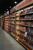 Shelves filled with various packaged rice and grains line a grocery store aisle. Brightly colored packaging and price tags are visible, with overhead signs in Spanish indicating the types of products available. Dim lighting adds a subdued atmosphere to the scene.