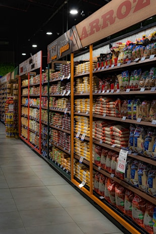 Shelves filled with various packaged rice and grains line a grocery store aisle. Brightly colored packaging and price tags are visible, with overhead signs in Spanish indicating the types of products available. Dim lighting adds a subdued atmosphere to the scene.