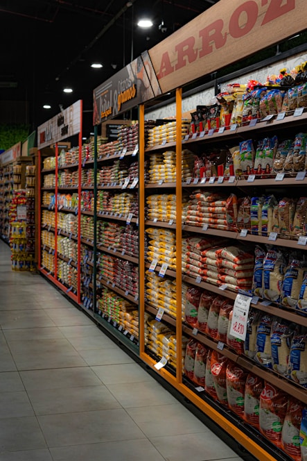 Shelves filled with various packaged rice and grains line a grocery store aisle. Brightly colored packaging and price tags are visible, with overhead signs in Spanish indicating the types of products available. Dim lighting adds a subdued atmosphere to the scene.