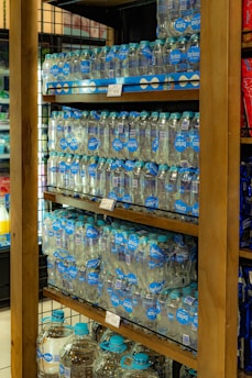 Shelves stocked with multiple packs of bottled water, organized neatly with clear labels displaying the brand 'brisa'. Each bottle has a blue cap and is part of a multi-pack held together by packaging. The shelves are wooden, and the setting appears to be in a retail environment with similar items displayed nearby.