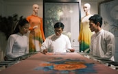 A group of three people is gathered around a table, focusing intently on an art piece or textile in a serene setting. Two mannequins dressed in traditional, colorful clothing are positioned in the background. The atmosphere suggests a blend of traditional craft and modern presentation.